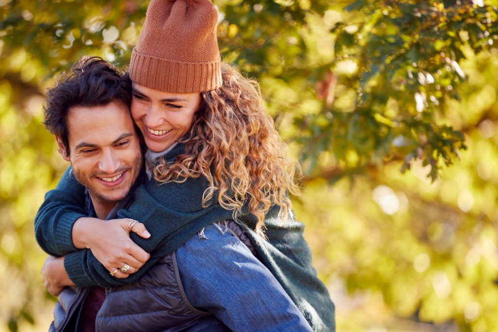 Happy Loving Couple Smiling As Man Give Woman Piggyback In Autumn Park